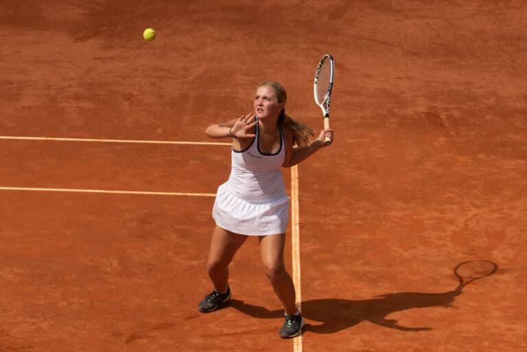 A female tennis player in a white outfit prepares to hit an overhead shot on a clay court, eyes focused on the ball above her, with her racket raised and shadow visible on the ground.