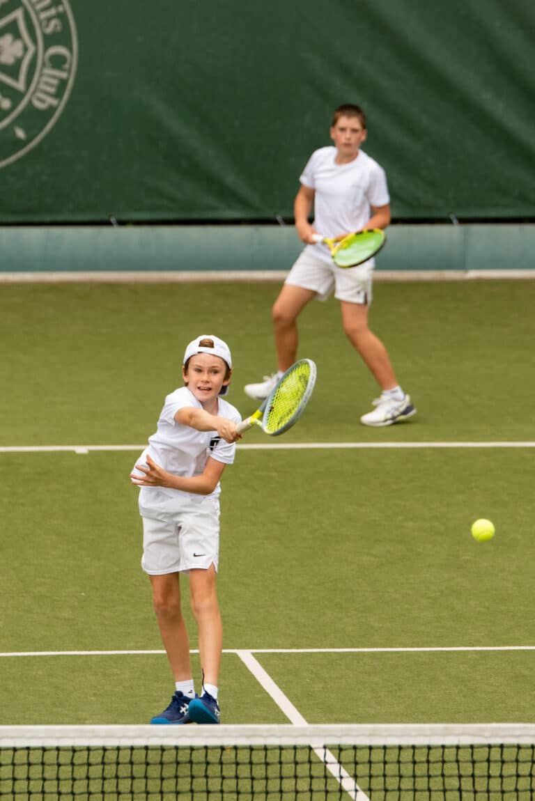 Two boys in white sportswear play doubles tennis on a grass court. The boy in front is swinging his racket to hit the ball, while the other boy stands ready behind him, watching the play.