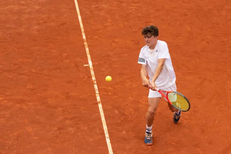 A male tennis player in a white outfit prepares to hit a backhand shot on a clay court. The yellow tennis ball approaches him, and he is focused on the action.