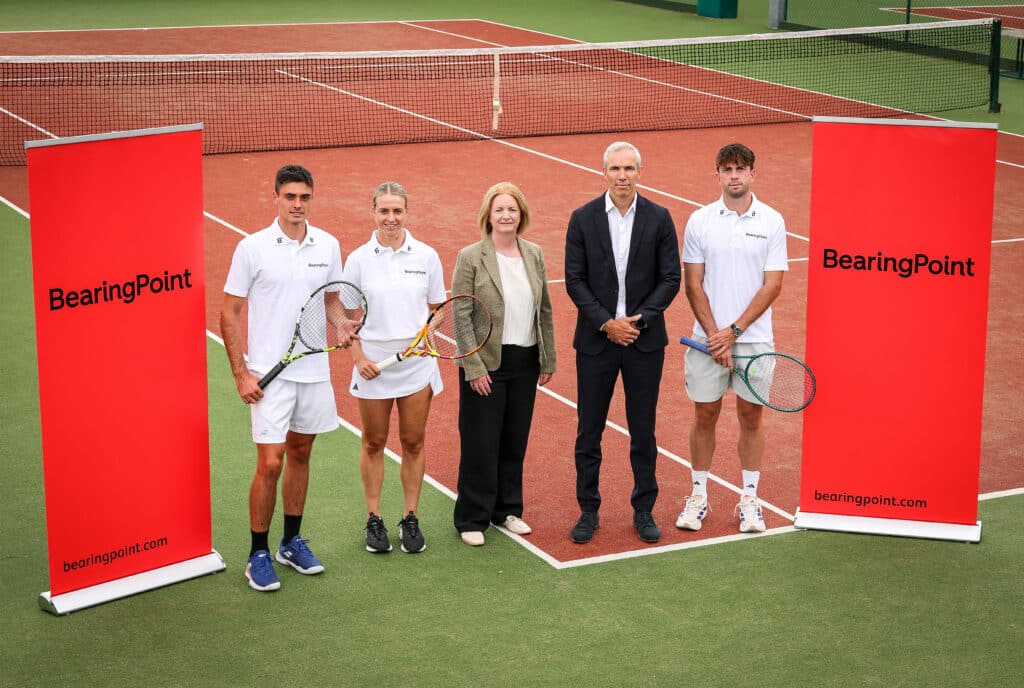 Five people standing on a tennis court with BearingPoint branding and three people are holding tennis rackets