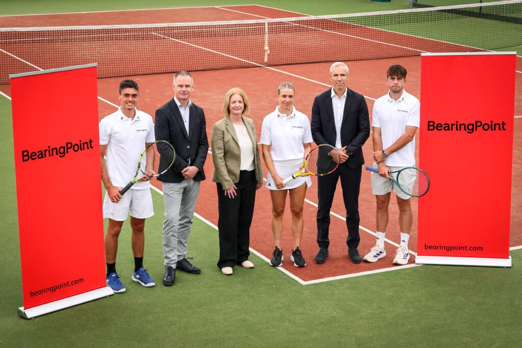 Six people standing on a tennis court with BearingPoint branding and three people are holding tennis rackets