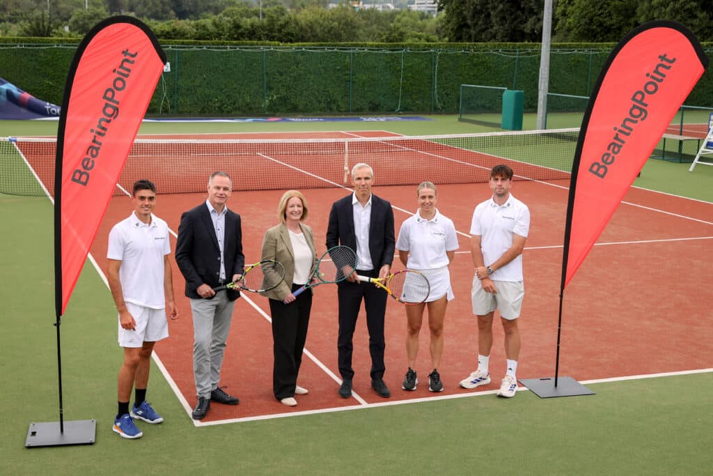 Six people standing on a tennis court with BearingPoint branding and three people are holding tennis rackets